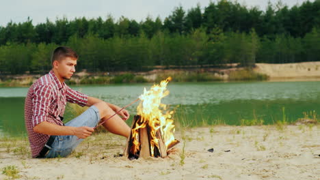 Junger-Attraktiver-Mann,-Der-Am-Lagerfeuer-Sitzt,-Braten-Marshmallows-Auf-Einem-Stock-Vor-Dem-Hintergrund-Von