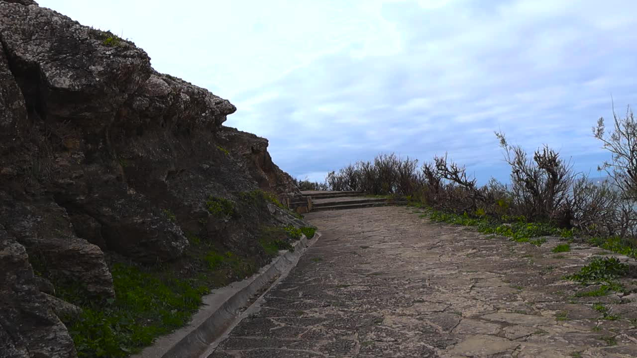Stabilized walking shot along a rocky clifftop path toward an ocean viewpoint, with rugged stone formations and open sky creating a calm coastal atmosphere