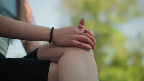 close-up of young woman seated outdoors with leg crossed, hands clasped together on knee, wearing black hand bangle, gently moving fingers, with blurred background featuring greenery