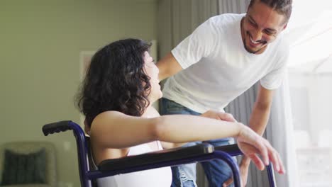 mujer biracial feliz en silla de ruedas y compañero masculino sonriente abrazándose en la sala de estar soleada