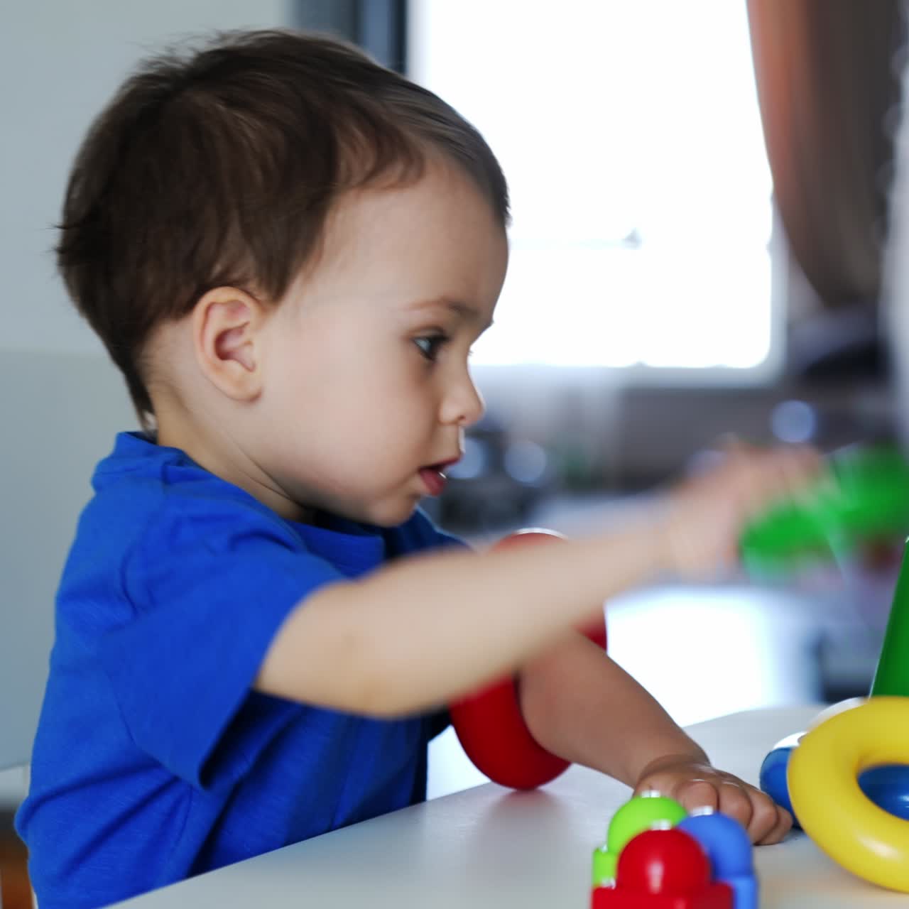 Un adorable bebé caucásico se sienta en el escritorio jugando con una pirámide. El niño pequeño se pone los coloridos anillos en las manos y aplaude alegremente.