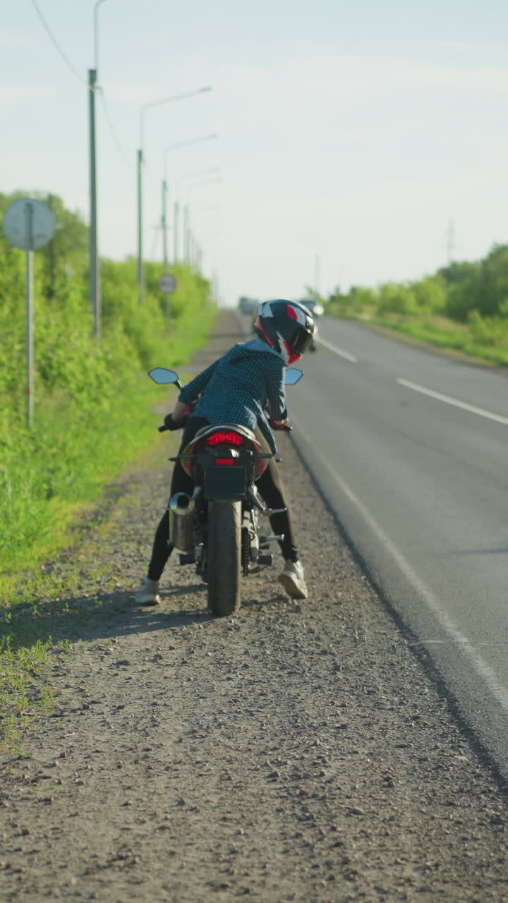 una mujer en una motocicleta mirando hacia atrás mientras comienza su bicicleta por la carretera, un arbusto verde exuberante bordea la carretera a medida que los coches se acercan desde la dirección opuesta, y la gente camina a lo largo de la carretera
