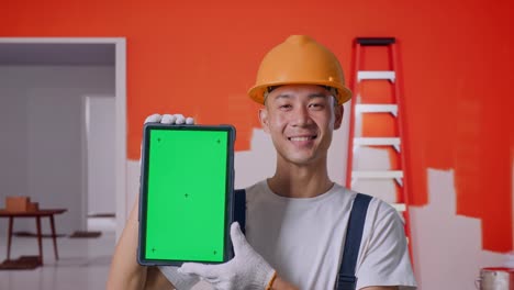 Close Up Of Asian Man Painter Wearing Safety Helmet Smiling And Showing Green Screen Tablet To The Camera While Standing With Painting Wall In Room