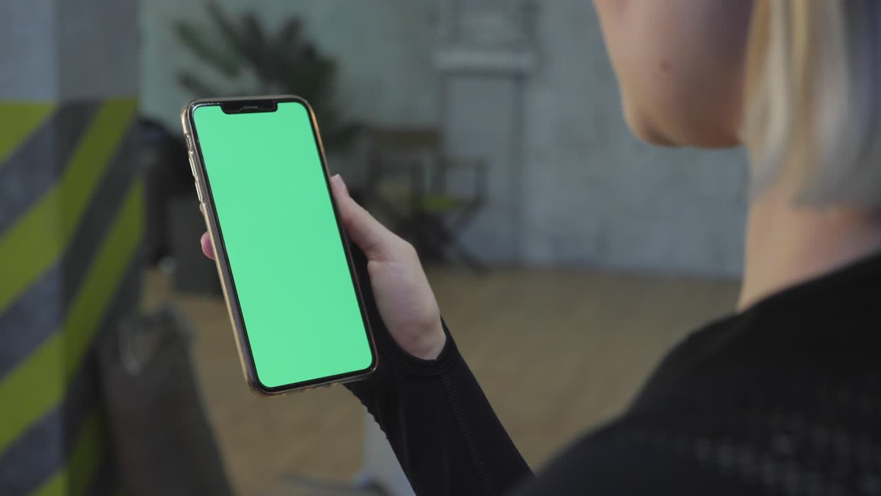 una joven atlética sostiene un teléfono inteligente con una pantalla verde chromakey en su mano, está en el gimnasio para entrenar