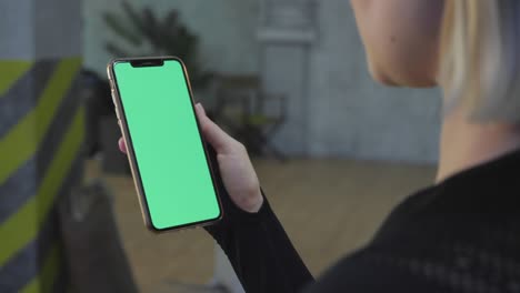 a young athletic girl holds a smartphone with a green chromakey screen in her hand, is in the gym for training