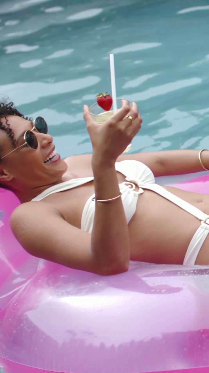 Vertical video: Lying lady in white bikini holding drink on pink float as friend in pool steadying