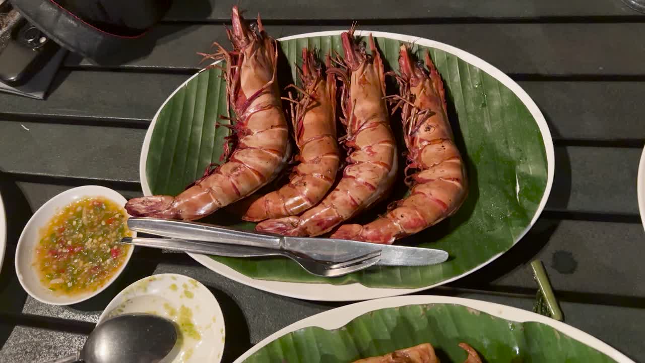 Hand removes grilled prawns from platter on outdoor dining table, evening lighting, close-up view