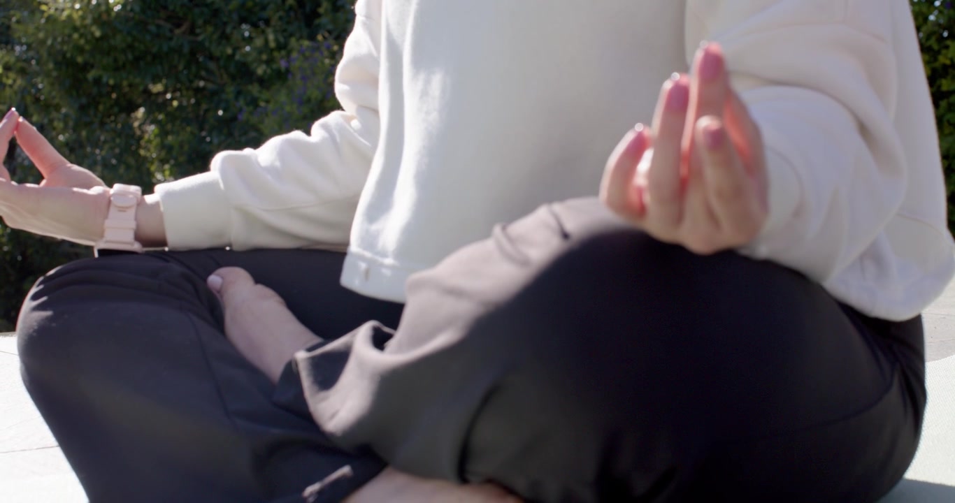 Kvinde justerer hofterne, sætter sig i skrædderstilling på en lys måtte nær en hæk, danner mudras for at meditere.