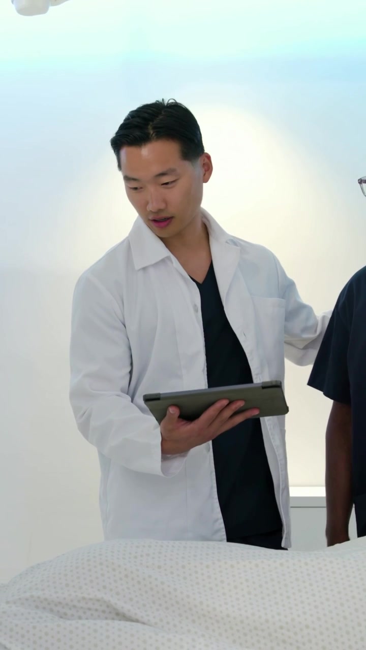 Video vertikal: Tablet berbunyi, dokter berjas putih meninjau pasien di meja pemeriksaan, merencanakan perawatan.
