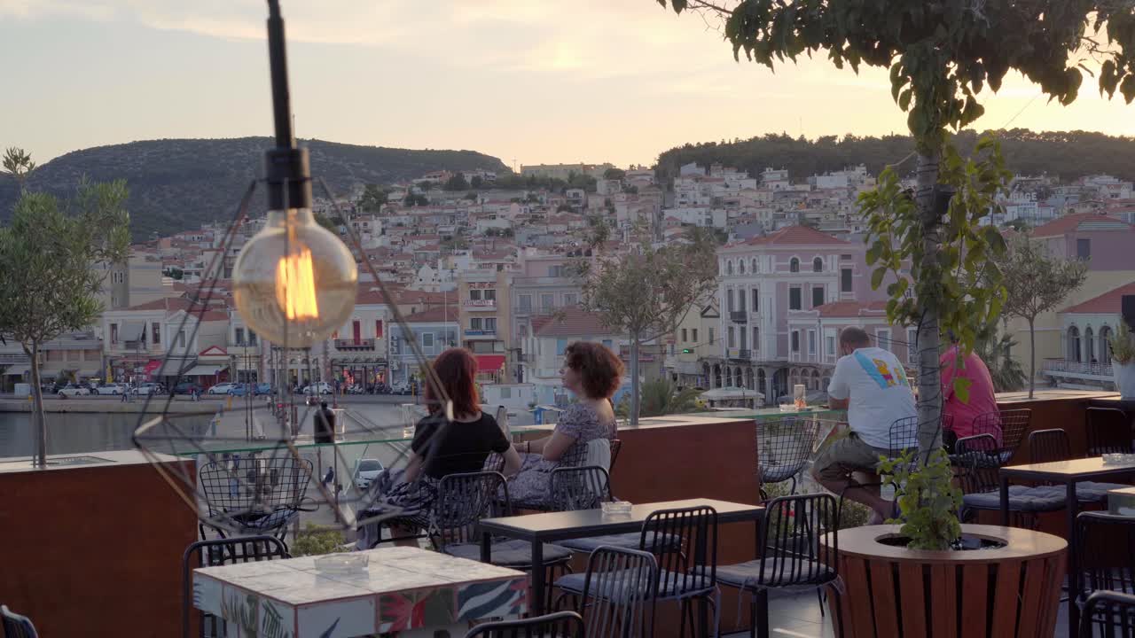 Women tourists sitting at a rooftop cafe overlooking the seafront and harbor of Mytilene city busy streets and buildings at sunset, Handheld gimbal shot