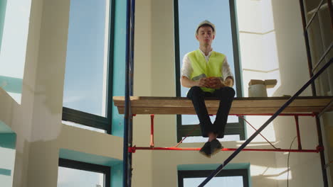 a worker eats a sandwich on the construction site, sits high on scaffolding