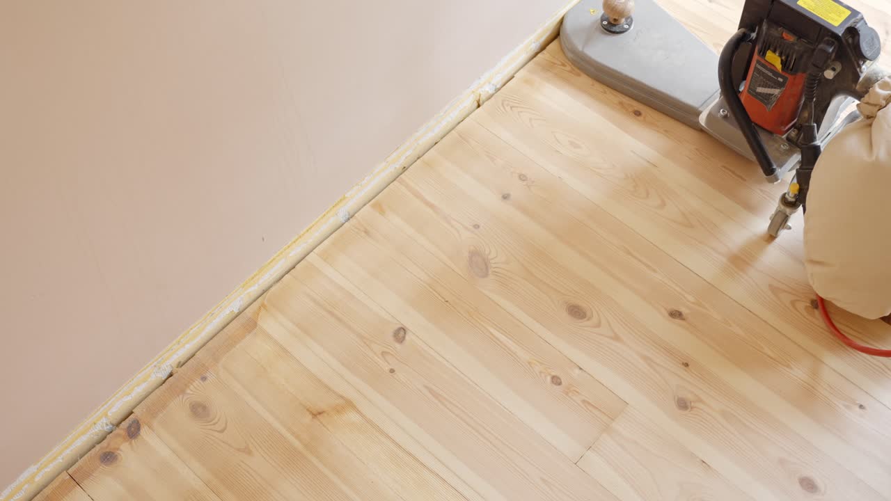 Using handheld sander to smooth wooden floor edge along interior wall, overhead view