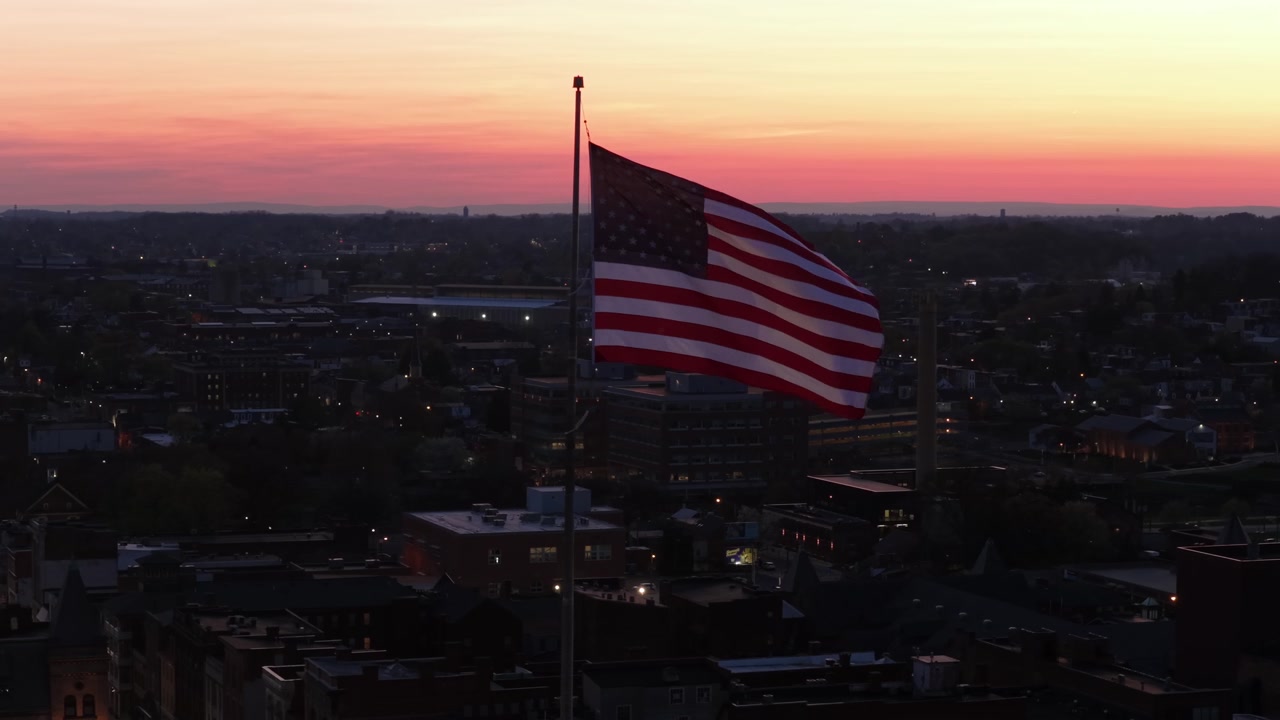 Bandiera americana che sventola su un evento patriottico al tramonto. Ripresa aerea in orbita. Città orgogliosa e pacifica negli USA. Edifici storici e centro città sullo sfondo. Strisce e stelle della bandiera, ripresa dettagliata.