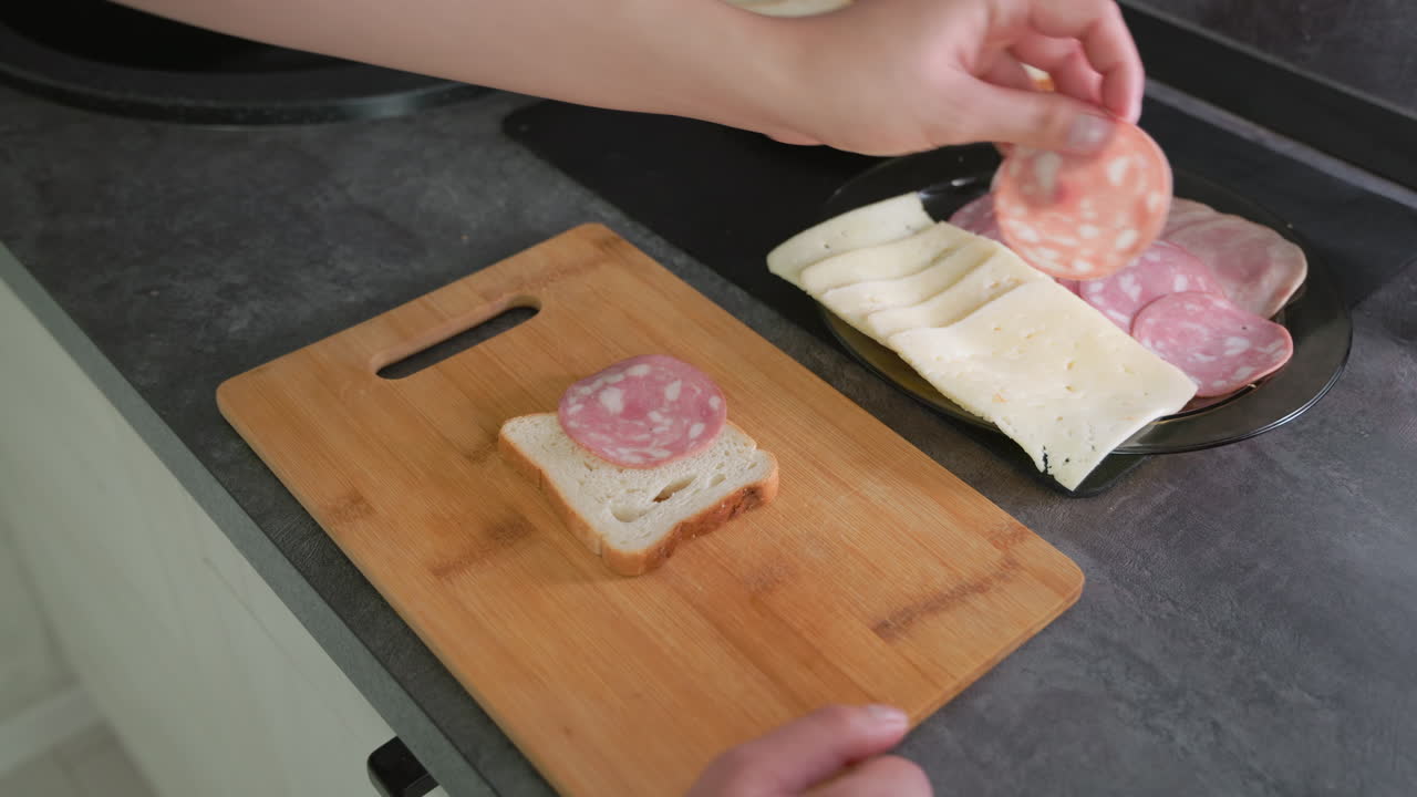 Hand selecting slice of salami from black plate with deli meats and cheese beside bread on wooden cutting board during sandwich preparation, showcasing casual home cooking routine in clean modern kitchen