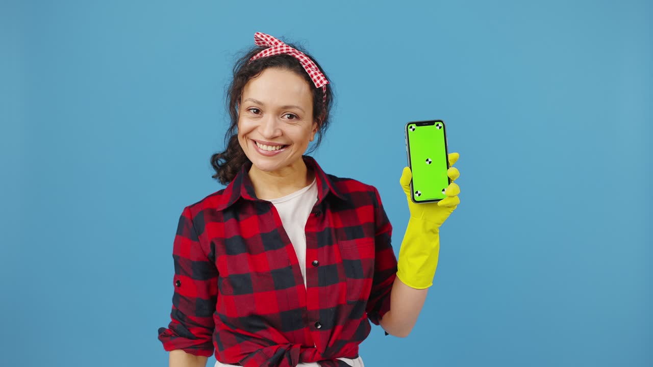Cheerful housewife in protective rubber gloves showing smartphone with chroma key screen for mockup, blue background