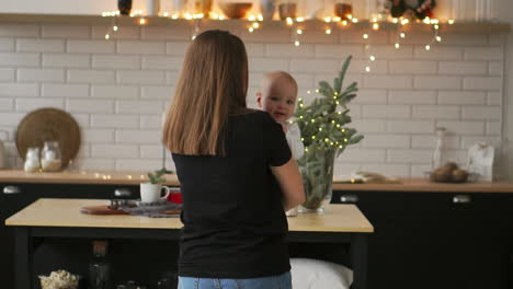 Mutter-Umarmt-Das-Baby-Und-Das-Kind-Lächelt-Und-Schaut-Seine-Geliebte-Mutter-An.-Gemeinsam-Stehen-Sie-Am-Heiligabend-In-Der-Weißen-Küche-Vor-Dem-Hintergrund-Von-Girlanden-Und-Weihnachtsbäumen.-Glückliche-Mutter-Und-Baby-Küssen-Und-Umarmen.-Mutterschaftskonzept-Mutterschaft-Schöne-Glückliche-Familie-Stockvideomaterial-In-Zeitlupe