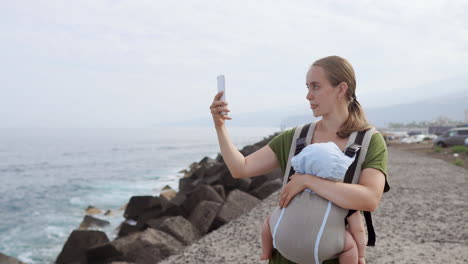 a lo largo de la orilla del océano, una madre y su bebé disfrutan de un sereno día de verano al aire libre. el niño encuentra consuelo en una mochila de canguro mientras caminan juntos