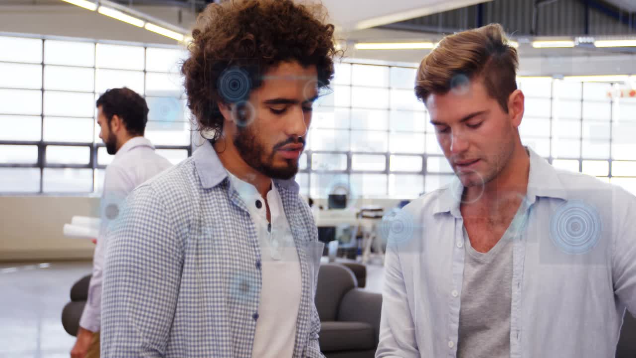 Two coworkers in tech office standing together, right man gesturing to palm and HUD overlay pulsing