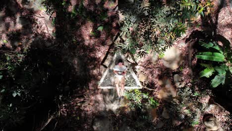 Lowering-aerial-of-a-young-traveler-laying-in-a-triangle-shaped-hammock-in-jungle-of-Colombia,-flying-his-drone,-sunny-day