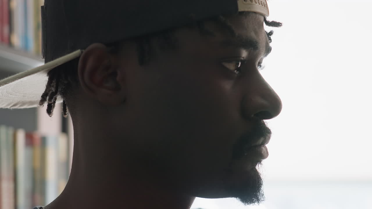 Extreme close up side profile of young man in cap with intense focused expression, eyes fixed ahead while books blur in background, conveying concentration