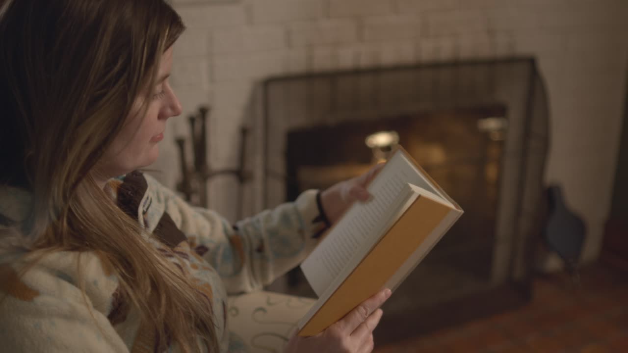 Over the shoulder shot of a woman in a sweater reading a book and flipping pages in front of a fire place. A relaxing, cozy, fall setting. Shot at a frame rate of 60 frames