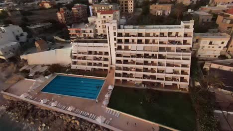 Vuelo-Aéreo-Hacia-Atrás-Desde-La-Ventana-Del-Complejo-Que-Revela-La-Playa-De-Batroun-Al-Atardecer,-Líbano