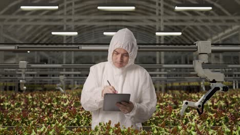 asian man researcher looking to side and taking note on a tablet while standing in the greenhouse with smart robotic farmers