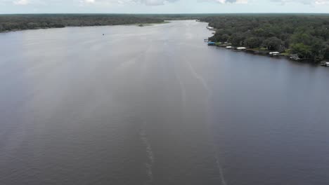 Aerial-view-overlooking-the-water-marina-speedboat-passing-by-in-water-sailboat-vessel-fisherman-greenery-and-trees-in-background