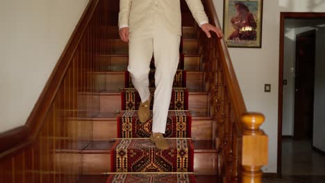 man in white suit descending a wooden staircase with decorative carpet runner