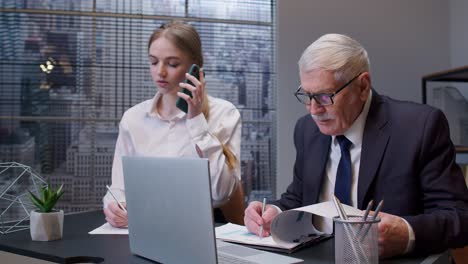 woman secretary talking on mobile cell phone, senior business man boss working on laptop in office