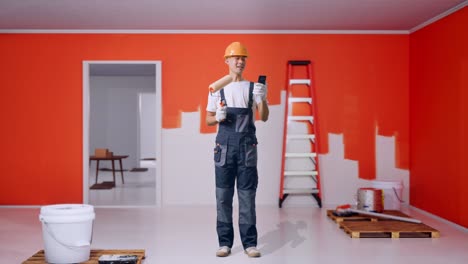 full body of asian man painter wearing safety helmet waving hand having a video call on smartphone while standing with painting wall in room