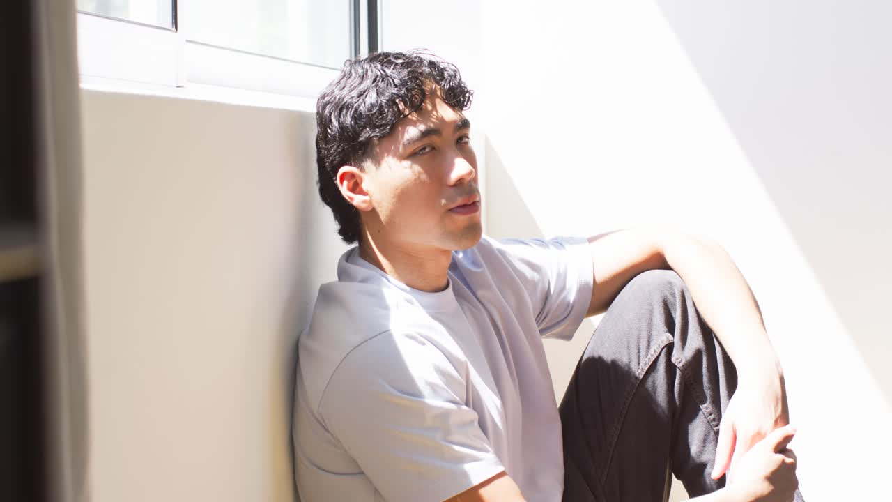 Man in his 20s sitting on floor by window, turning toward camera wearing light gray t-shirt