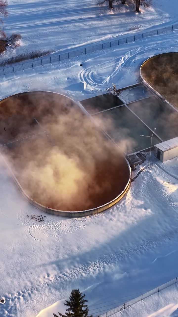 Flygfoto över ångande vattenreningsbehållare i ett snötäckt industriområde.
