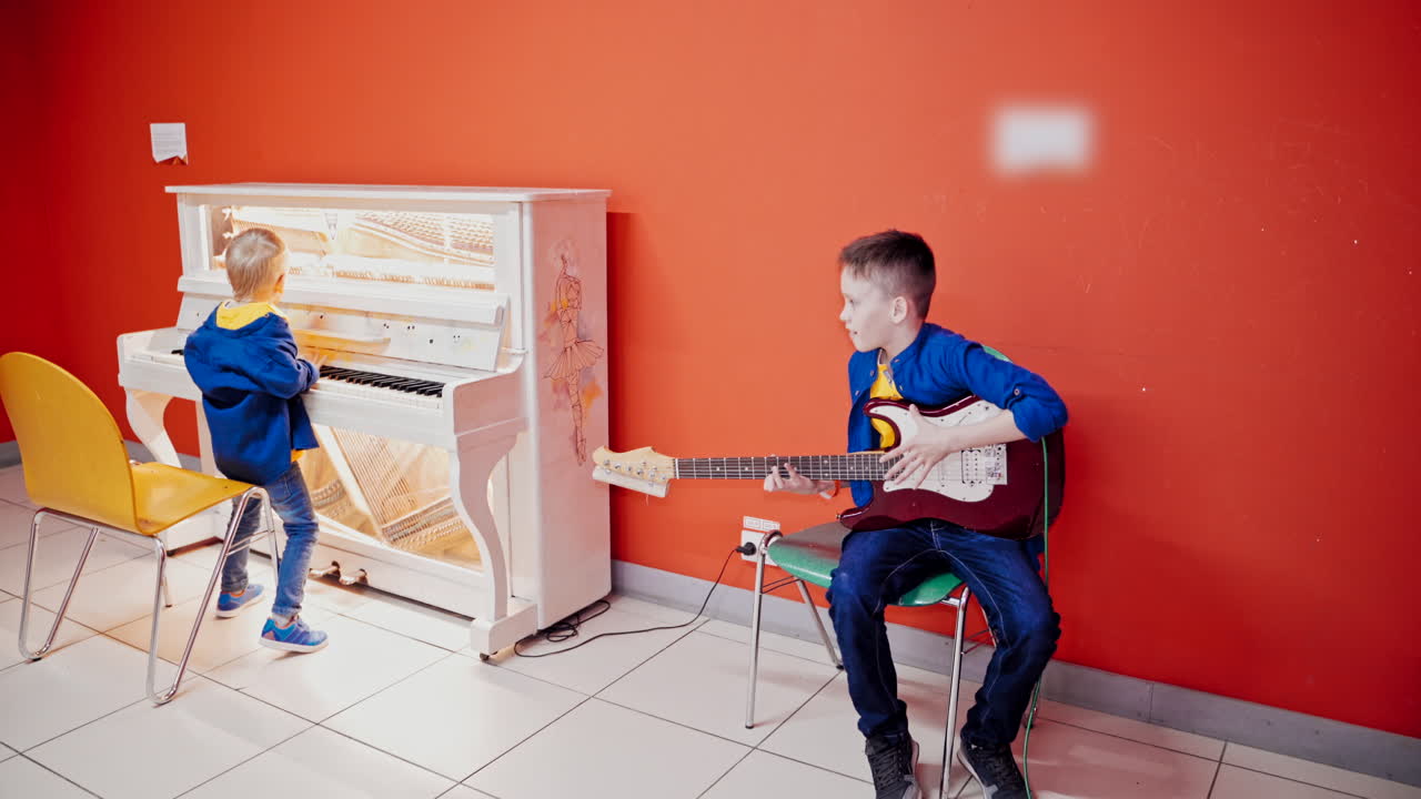 Two happy boys are playing on musical instruments on the red wall background. One boy is playing on the acoustic guitar and another smaller boy is playing the piano.