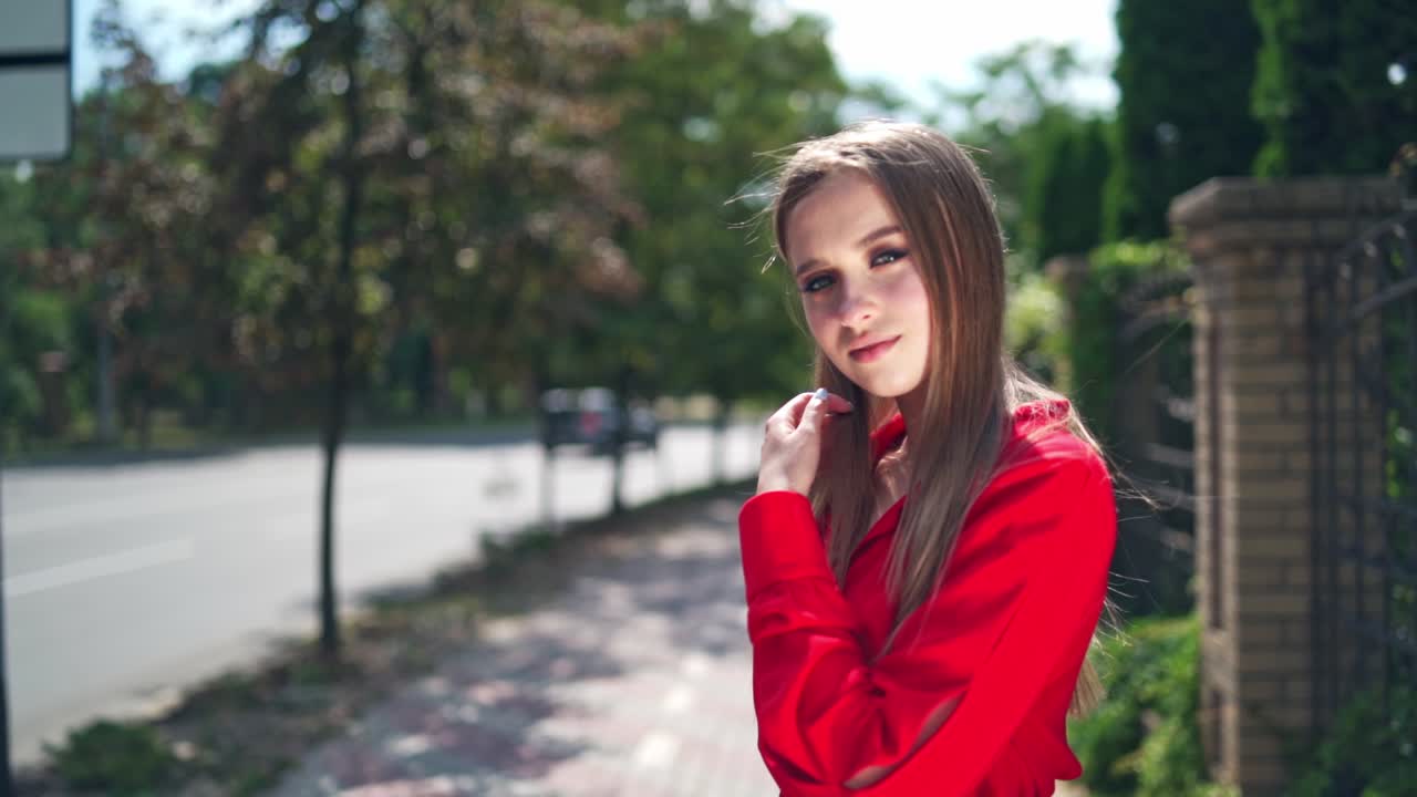 Lovely girl in red dress. Beautiful young model is standing outdoors and posing on camera. Portrait of a pretty woman on the green trees background.