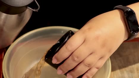 a person pours dark liquid from a black cup into a transparent bowl on a wooden surface.