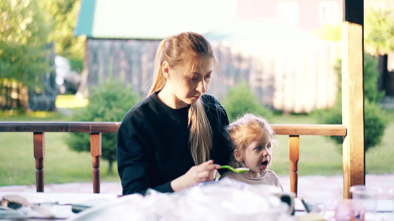 예쁜 젊은 어머니는 딸에게 가루를 먹이고 있습니다. 그들은 여름 집에 앉아 있습니다.