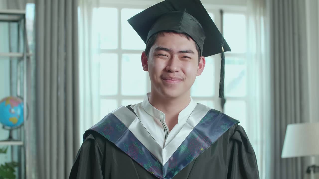 卒業式のドレスとキャップを着たアジア人男性がリビングでカメラに微笑んでいる