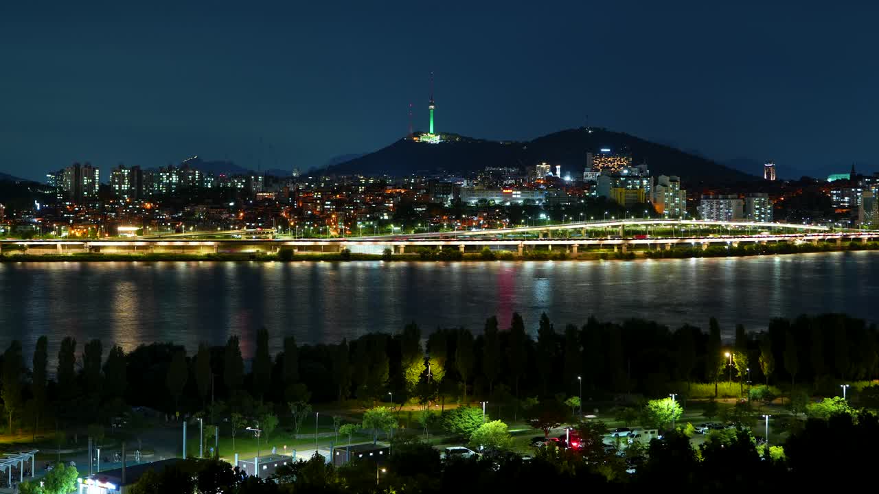 A beautiful time-lapse of the iconic N Seoul Tower and the city skyline at night, with the vibrant lights of a riverside highway and buildings reflecting beautifully in the tranquil Han River
