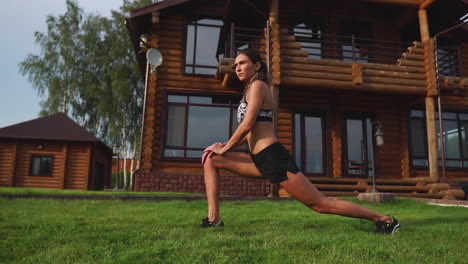 A-slim-and-beautiful-woman-in-sportswear-with-an-open-press-is-preparing-to-start-training-on-the-lawn-near-her-home-flexing-her-legs,-knees,-shins-and-thighs-with-warming-up-exercises