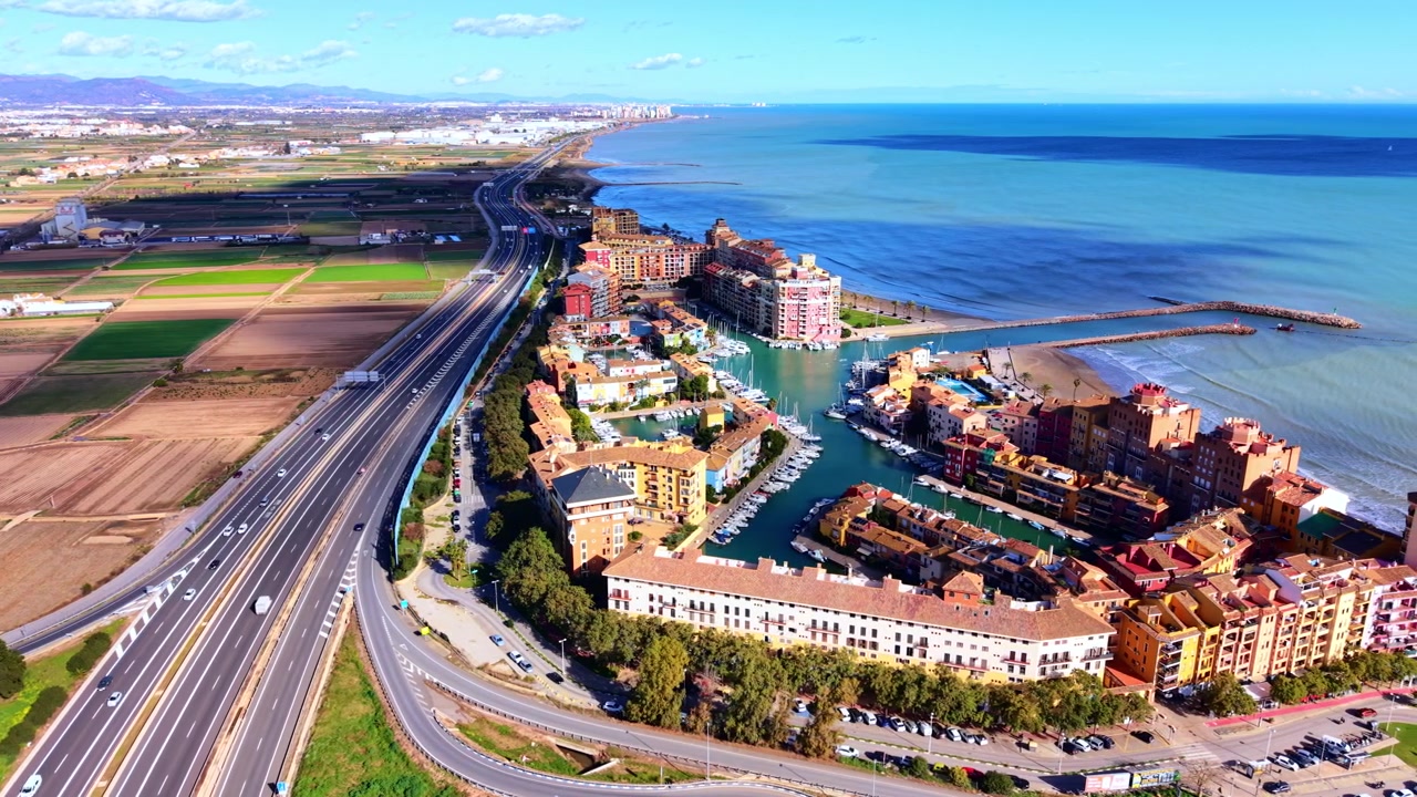 Autostrada costiera e resort con marina, Castellón, Spagna. Prospettiva panoramica di strada a più corsie, terreni agricoli, Mar Mediterraneo, spiaggia sabbiosa ed edifici colorati sul lungomare.