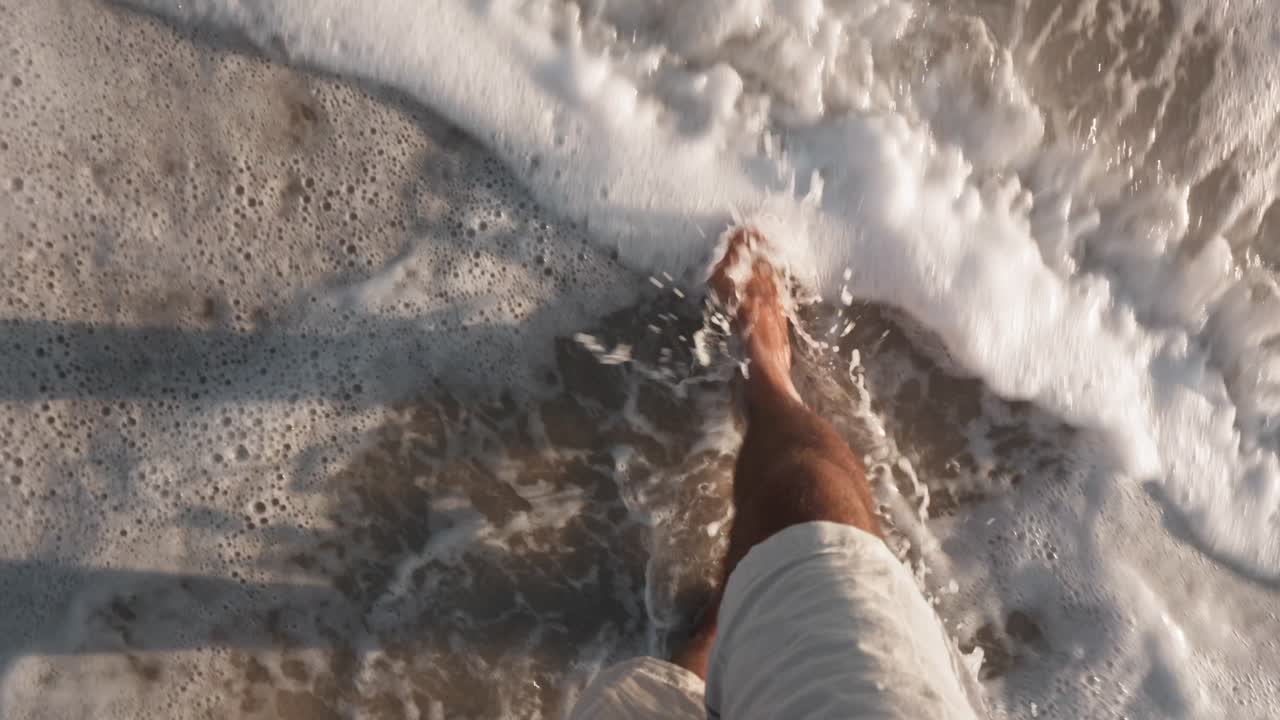POV shot capturing a man’s barefoot footsteps walking along a sandy coastline. The camera follows the steps, showcasing the texture of the sand and the gentle lapping of waves in the background.
