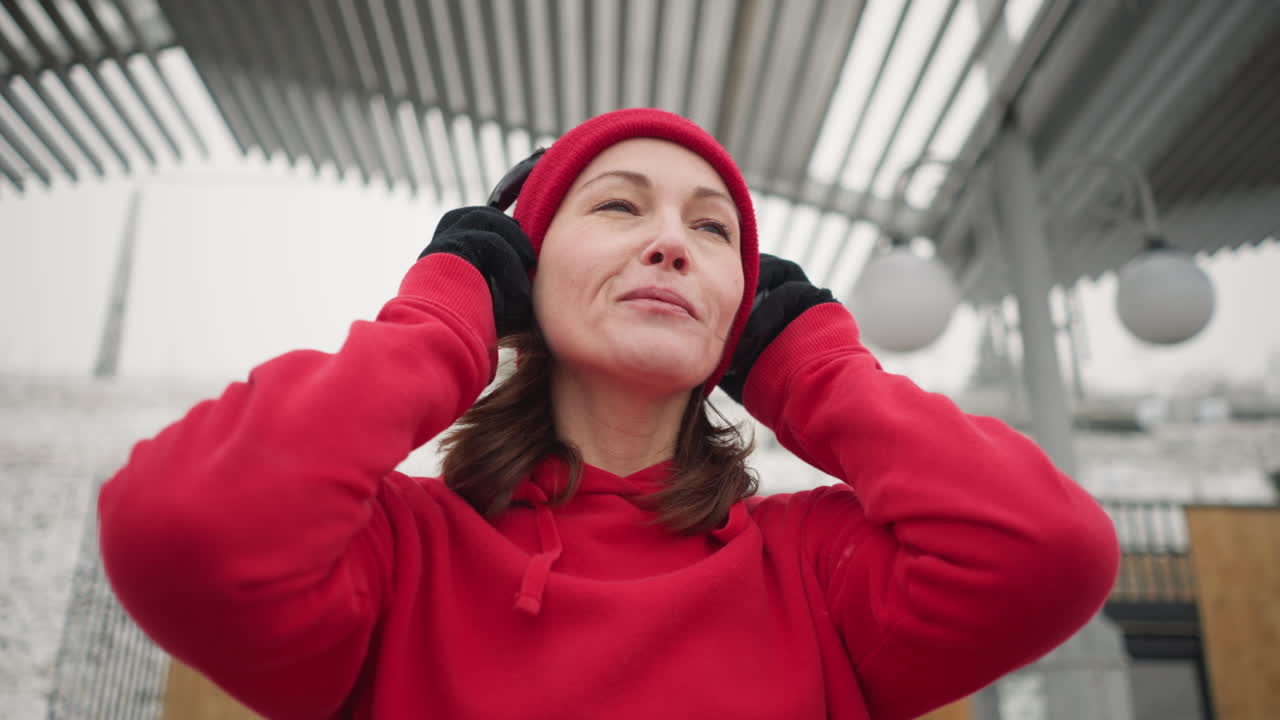 mujer con gorra roja y sudadera con capucha sonríe mientras ajusta los auriculares al aire libre en invierno, el fondo presenta un paisaje urbano cubierto de nieve, arquitectura moderna y una atmósfera de niebla serena