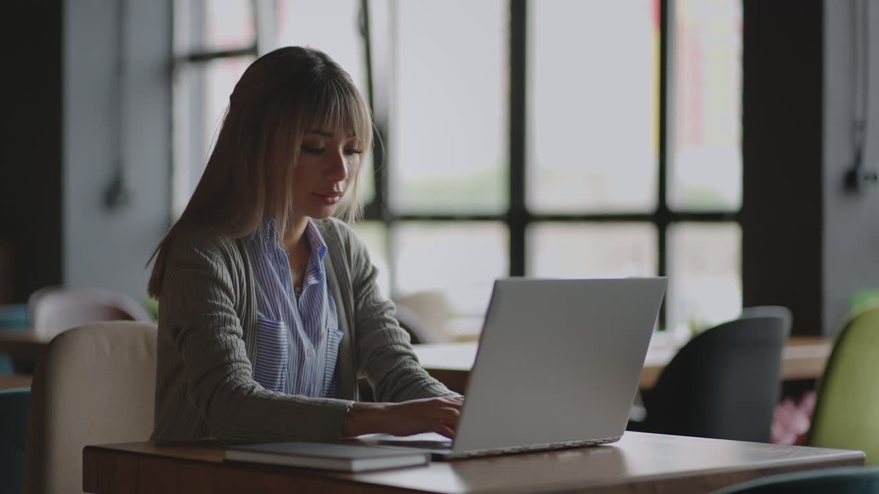 Asian lady is using laptop at university library typing then taking notes in notebook.