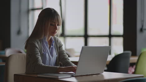 Una-Mujer-Asiática-Está-Usando-Una-Computadora-Portátil-En-La-Biblioteca-De-La-Universidad-Escribiendo-Y-Luego-Tomando-Notas-En-Un-Cuaderno.