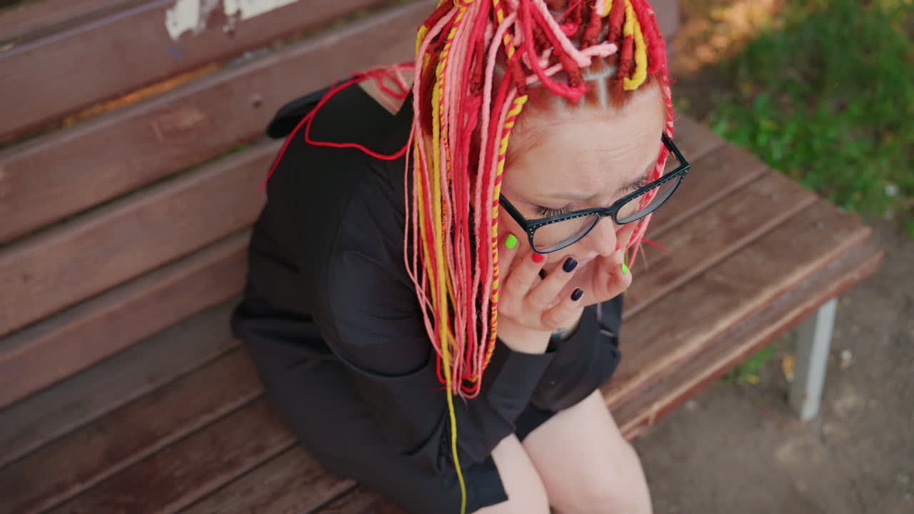 primer plano de una mujer blanca sentada en un banco tapándose la boca con las uñas pintadas, trenzas de colores y gafas, expresión intensa de preocupación, listones del banco del parque y vegetación suave de fondo, momento emocional crudo