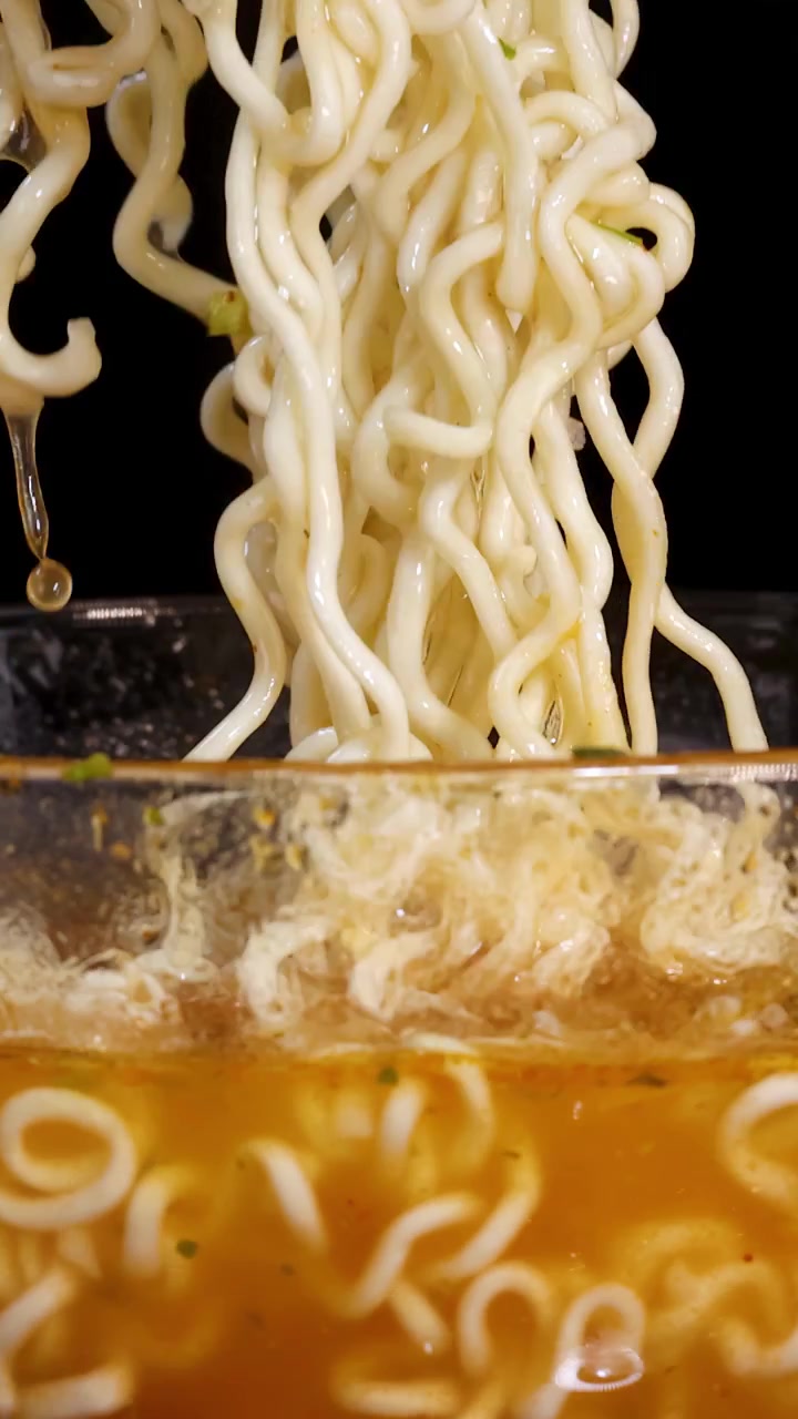 Una mano solleva noodles istantanei cotti da una ciotola di vetro fumante di brodo, con primi piani drammatici, illuminazione vibrante e uno sfondo scuro