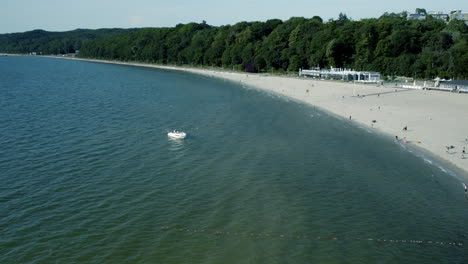 aerial: white boat anchored on shoreline of sea with sandy beach and forest trees in background - marina in gdynia, poland