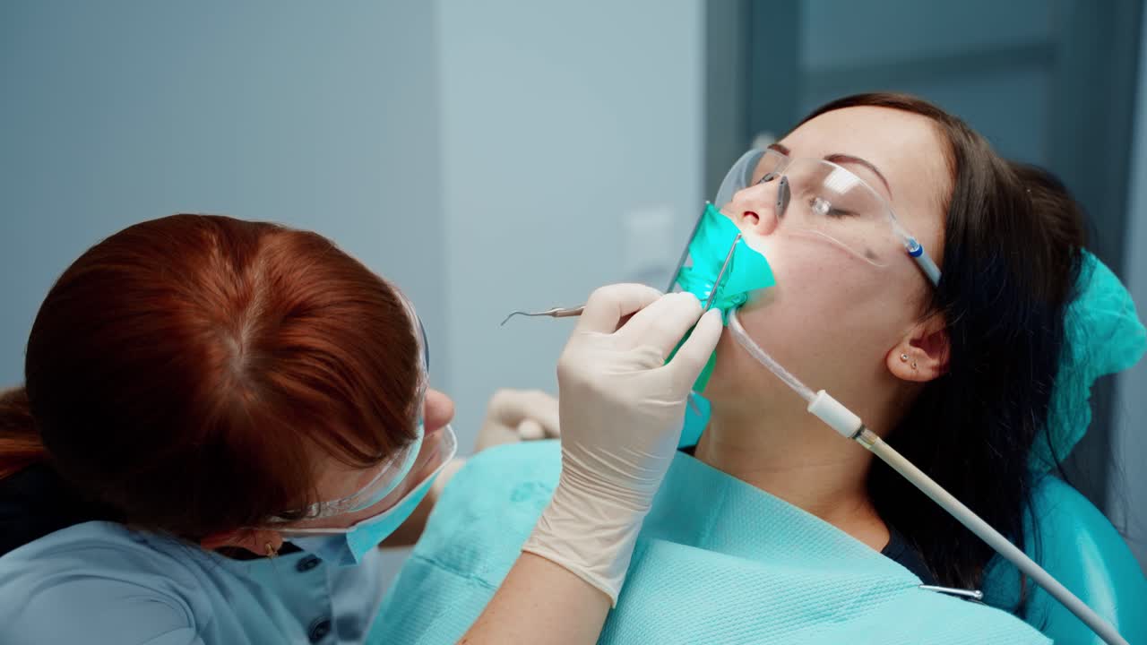 Woman in chair at stomatology clinic. Professional female dentist in protective mask and glasses treating patient's teeth using medical equipment.