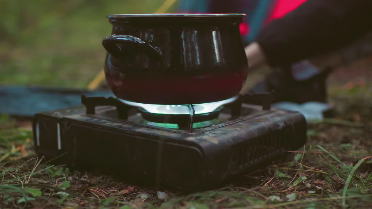 close up of black metal pot on portable gas stove with visible flame as person adjusts burner while cooking outdoors on grassy forest ground surrounded by leaves and scattered camping gear